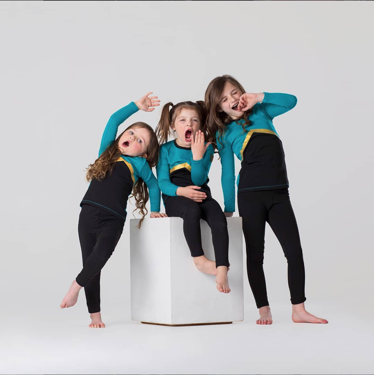 Three girls in teal dance costumes striking dramatic poses around a white cube