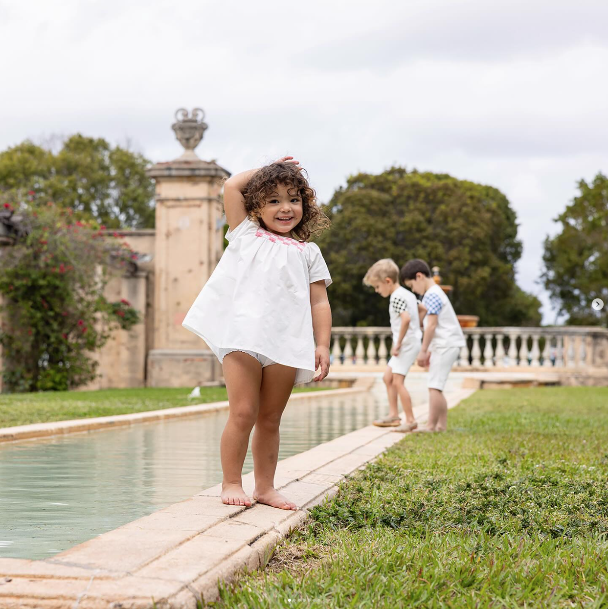 A smiling girl in a white dress beside a garden pool, with two children behind her.