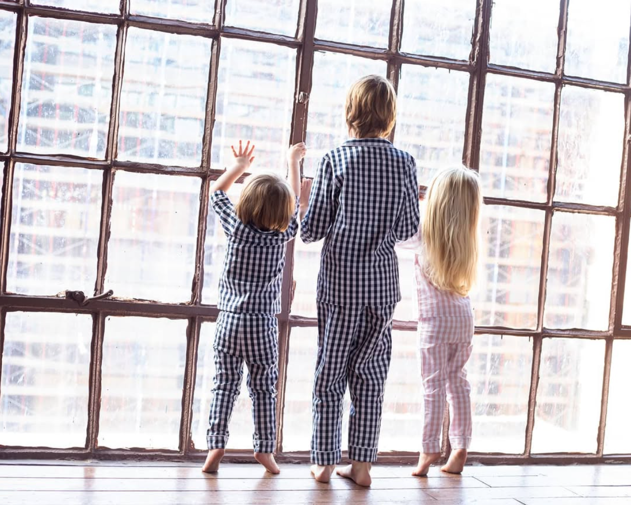 Three children in checked pajamas standing in the light of a large studio window.