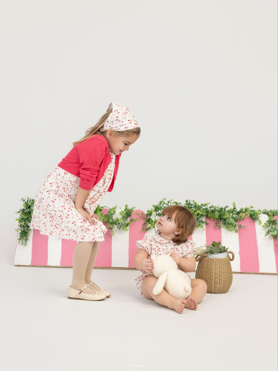 Two girls in floral outfits looking at each other beside a pink-striped planter, the younger holding a plush bunny.