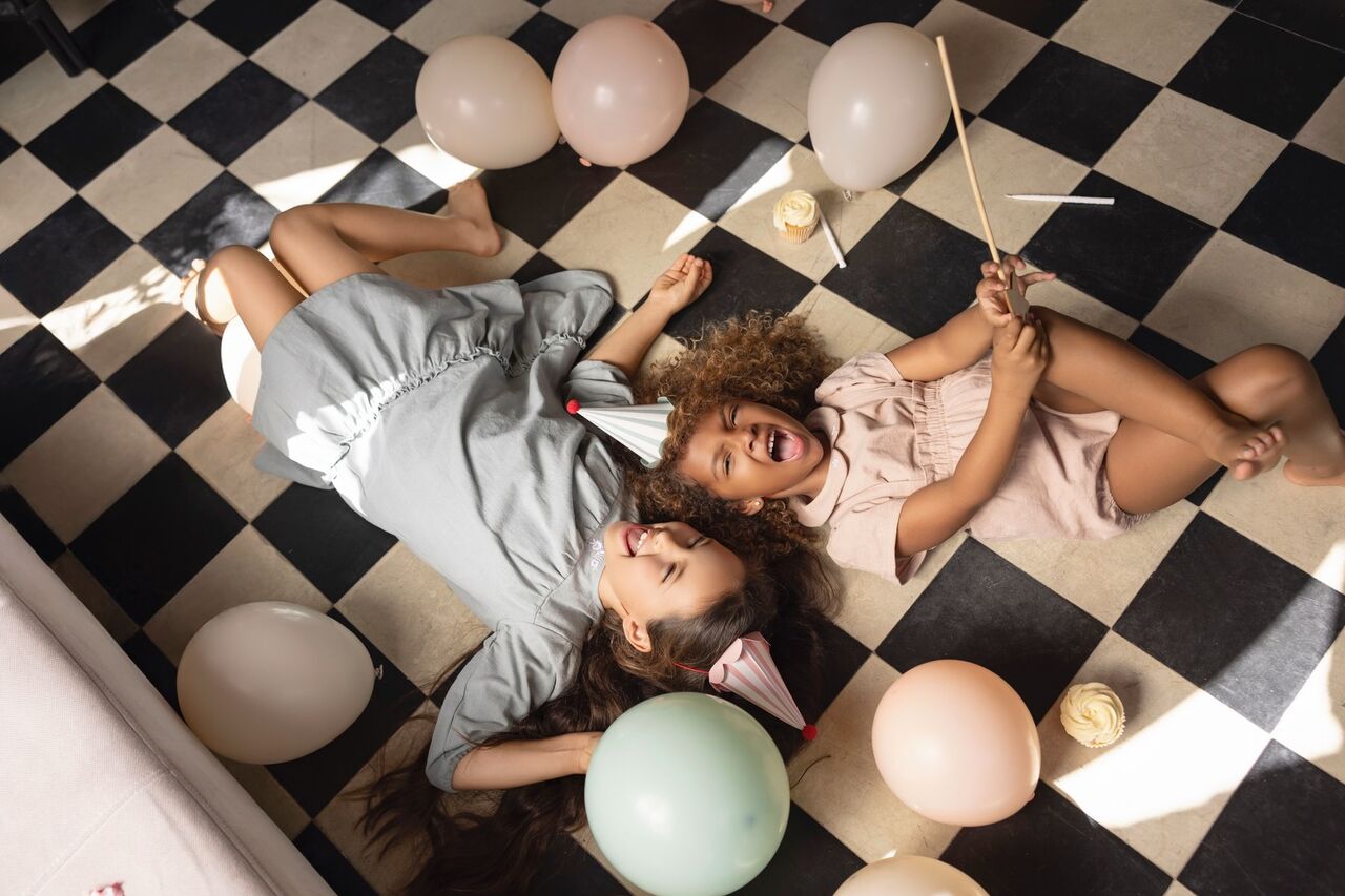 Two girls in party hats lying on a checkered floor among balloons