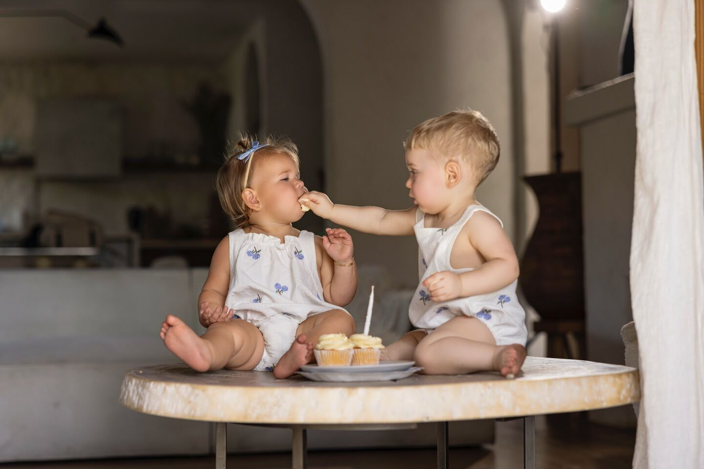 Two toddlers sitting on a wooden table, one feeding the other a cupcake